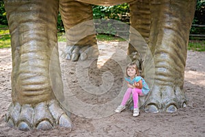 Little girl sitting under huge Diplodocus dinosaur sculpture