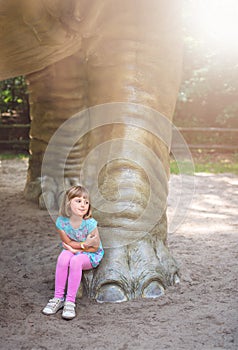 Little girl sitting under huge Diplodocus dinosaur sculpture