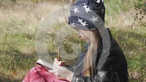 Little girl sitting under the tree and drawing in notebook at park. Beautiful little girl draws outdoors lifestyle