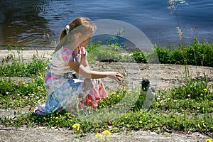 A little girl is sitting by the water and reaches out to a small crow