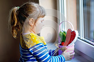 Little girl sitting by window with hyacinth flowers and hand-made heart. Happy child, indoors. Mother& x27;s day