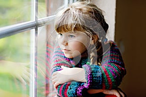 Little girl sitting by window. Preschool child wear cozy self knitted wool sweater. Toddler watching on rain outside