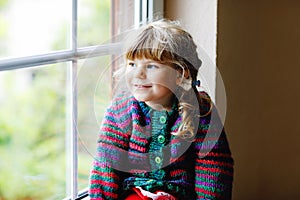 Little girl sitting by window. Preschool child wear cozy self knitted wool sweater. Toddler watching on rain outside