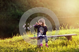 Little girl sitting on a wooden bench blows bubbles in the rays