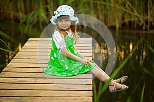 Little girl sitting on wooden bridge across river in summer day