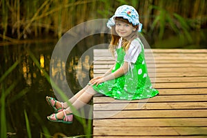 Little girl sitting on wooden bridge across river in summer day