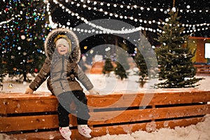 Little girl is skating on ice skates on skating rink in evening, decorated with fairy lights, Christmas trees and fir. Festive