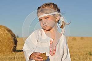 A little girl in slavic traditional ornamented chemise eating rye bread in the harvested filed