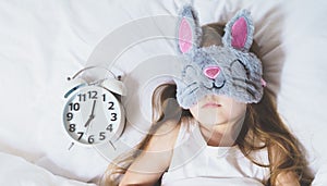 Little girl sleeping in bed under white blanket wearing grey bunny plush sleep mask with alarm clock on pillow. Early morning wake