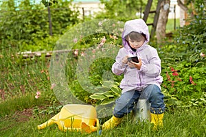 A little girl with a smartphone in her hand in the garden near a picturesque flower bed with hostas.
