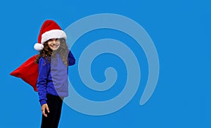 Little girl with smling wearing santa hat carrying gift bag isolated on blue background. Winter holidays. Copy space