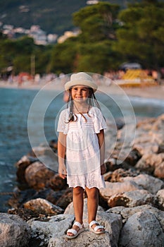 Little girl stand on rocks by the sea. Beautiful girl in white dress and hat
