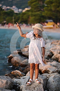 Little girl stand on rocks by the sea. Beautiful girl in white dress and hat