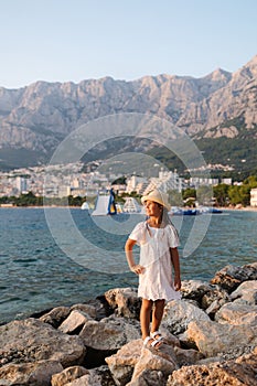 Little girl stand on rocks by the sea. Beautiful girl in white dress and hat