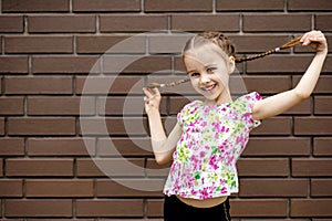 A little girl stands against a brick wall and playfully holds her pigtails