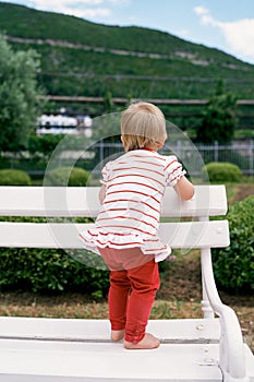 Little girl stands barefoot on a white bench in a green park. Back view