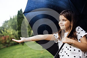 A little girl stands with a black umbrella in the rain. A cute baby stretches out her hand and catches raindrops in the park