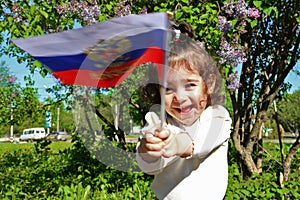 Little girl stands with flag of Russia in front of lilac Bush on sunny day