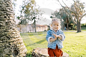 Little girl stands near a tree in a sunny meadow and cunningly rubs her hands