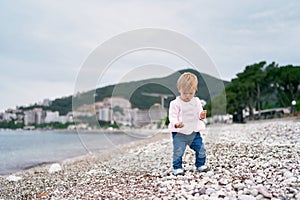 Little girl stands on a pebble beach and holds a pebble in her hand
