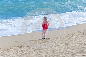 A little girl stands on a sandy beach and looks at the waves of the sea.