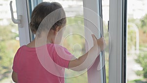 A little girl stands on the windowsill near the window of a high-rise building and holds on to the half-open window with