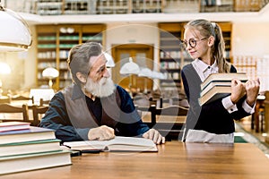 A little girl student pupil holding a lot of books in her hands and looking at her senior bearded teacher sitting at the