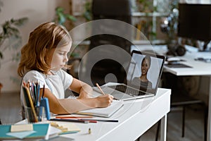 Little girl studies distantly at videocall via laptop at table in light room
