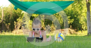 little girl in sunglasess is using laptop and lying on grass on summer sunny day. hammock and ball on the background