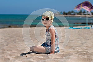 Little girl in sunglasses pours sand from her hands on the beach against the background of the sea