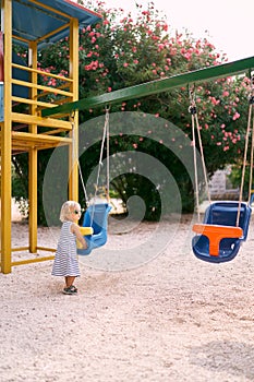 Little girl in sunglasses stands near the swing on the playground