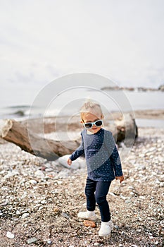 Little girl in sunglasses stands on a pebble beach near a driftwood