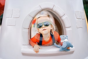 Little girl in sunglasses with a toy in her hand looks out of the window of a toy castle on the playground