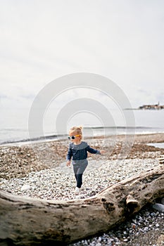 Little girl in sunglasses walks along the beach near a driftwood