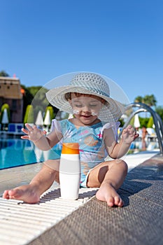 Cute baby girl looking at bottle of sunblock cream. Resort swimming pool in the background. Children summer vacation concept