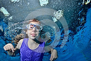 A little girl in swimming glasses is swimming underwater in the pool on a blue background and looking at me. Portrait. Close up. B