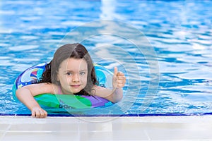 A little girl is swimming in the pool with inflatable ring and showing thumb`s up gesture. Kids learn to swim