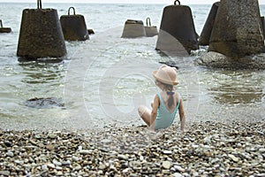 A little girl in a swimsuit and a hat runs on the seashore. The child cannot swim due to the breakwaters in the sea