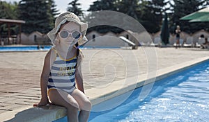 A little girl in a swimsuit and a hat sits near the pool with her feet in the water. Rest in warm countries. Rest at the hotel