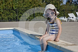 A little girl in a swimsuit and a hat sits near the pool with her feet in the water. Rest in warm countries. Rest at the hotel