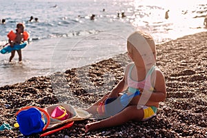 Little girl in a swimsuit sits by the sea and plays with sand toys in the summer in the contour light of the sun