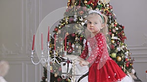A little girl is swinging while sitting on a toy horse in a room with a Christmas decor and a decorated Christmas tree.