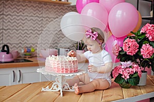 A little girl on the table with a birthday cake and balloons. The collapse of the cake. First birthday of the baby. Cakesmash