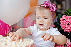 A little girl on the table with a birthday cake and balloons. The collapse of the cake. First birthday of the baby. Cakesmash