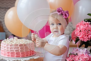 A little girl on the table with a birthday cake and balloons. The collapse of the cake. First birthday of the baby. Cakesmash