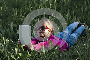 A little girl takes selfies on her tablet while lying in the green grass.