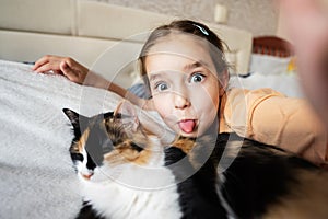 Little girl taking selfie with cat on bed at home. Portrait of adorable child show tongue with pet