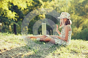 Little girl is talking on a laptop while sitting on the grass in the sun. Dressed in a sarafan and hat