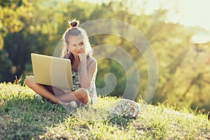 Little girl is talking on a laptop while sitting on the grass in the sun. Dressed in a sarafan and hat