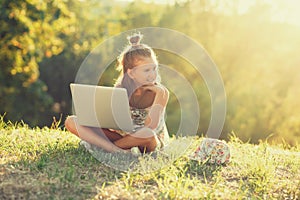 Little girl is talking on a laptop while sitting on the grass in the sun. Dressed in a sarafan and hat